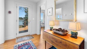 A hallway with a wooden console table, a mirror, and a door leading to a balcony. - Chimerique on the Water in Maine (Saint George)