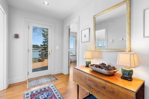 A hallway with a wooden console table, a mirror, and a door leading to a balcony. - Chimerique on the Water in Maine (Saint George)