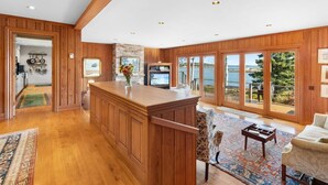 A spacious living area with a wooden bar, large windows with a view of the water, and a cozy seating area. - Chimerique on the Water in Maine (Saint George)