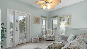 A living room with a beige sofa, a patterned armchair, a ceiling fan, and a door leading to a balcony. - Emerald Mist (Miramar Beach)