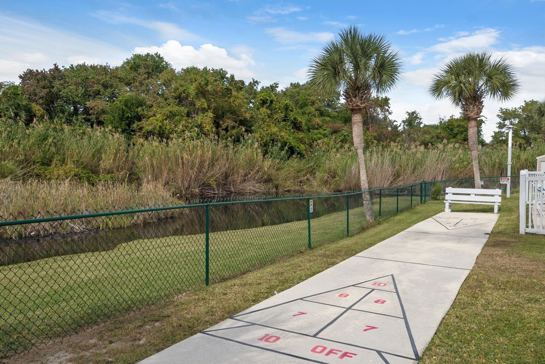 A shuffleboard court with a green fence, palm trees, and a bench.