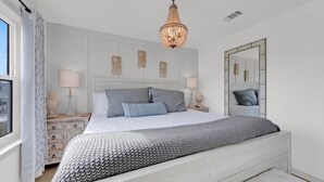 A bedroom with a bed, bedside tables, a chandelier, a mirror, and artwork on the wall. - Beachside Villas 1123- A Sandy Carol (Santa Rosa Beach)