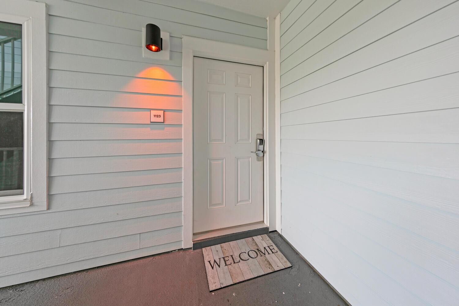 A white door with a keypad lock, a welcome mat, and a wall-mounted light fixture.