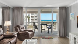 A spacious living room with a leather sofa, a glass coffee table, and a view of the beach through large sliding doors. - Shoreline Towers 2023 (Destin)
