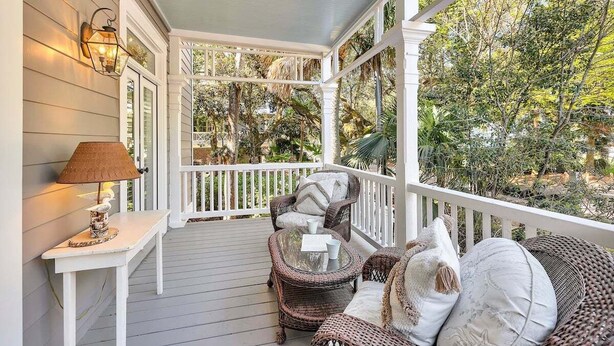 A covered porch with wicker furniture, a table with a lamp, and a view of trees and foliage. - Ebb Tide, Steps from the Beach & Community Pool (Saint Simons Island)