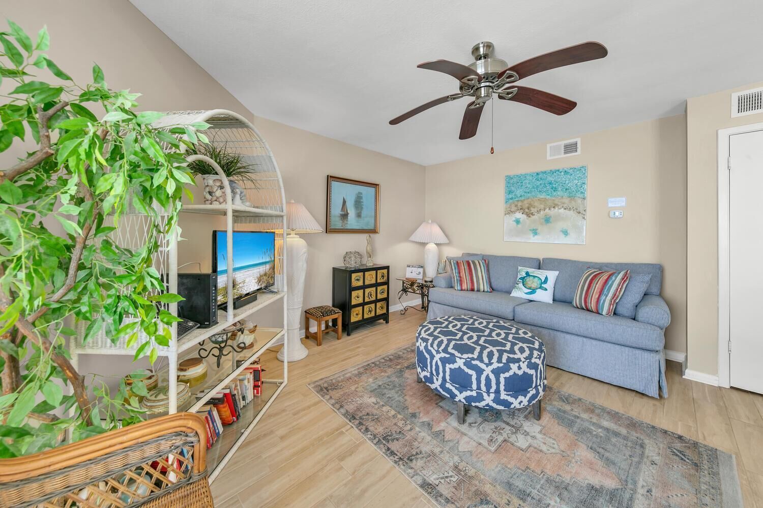A living room with a ceiling fan, a sofa, a patterned ottoman, a television on a white shelf, and a wall with a beach painting.