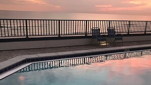 A swimming pool with a view of the ocean at sunset, a black metal fence, and two lounge chairs. - Aquavista 504w (Panama City Beach)