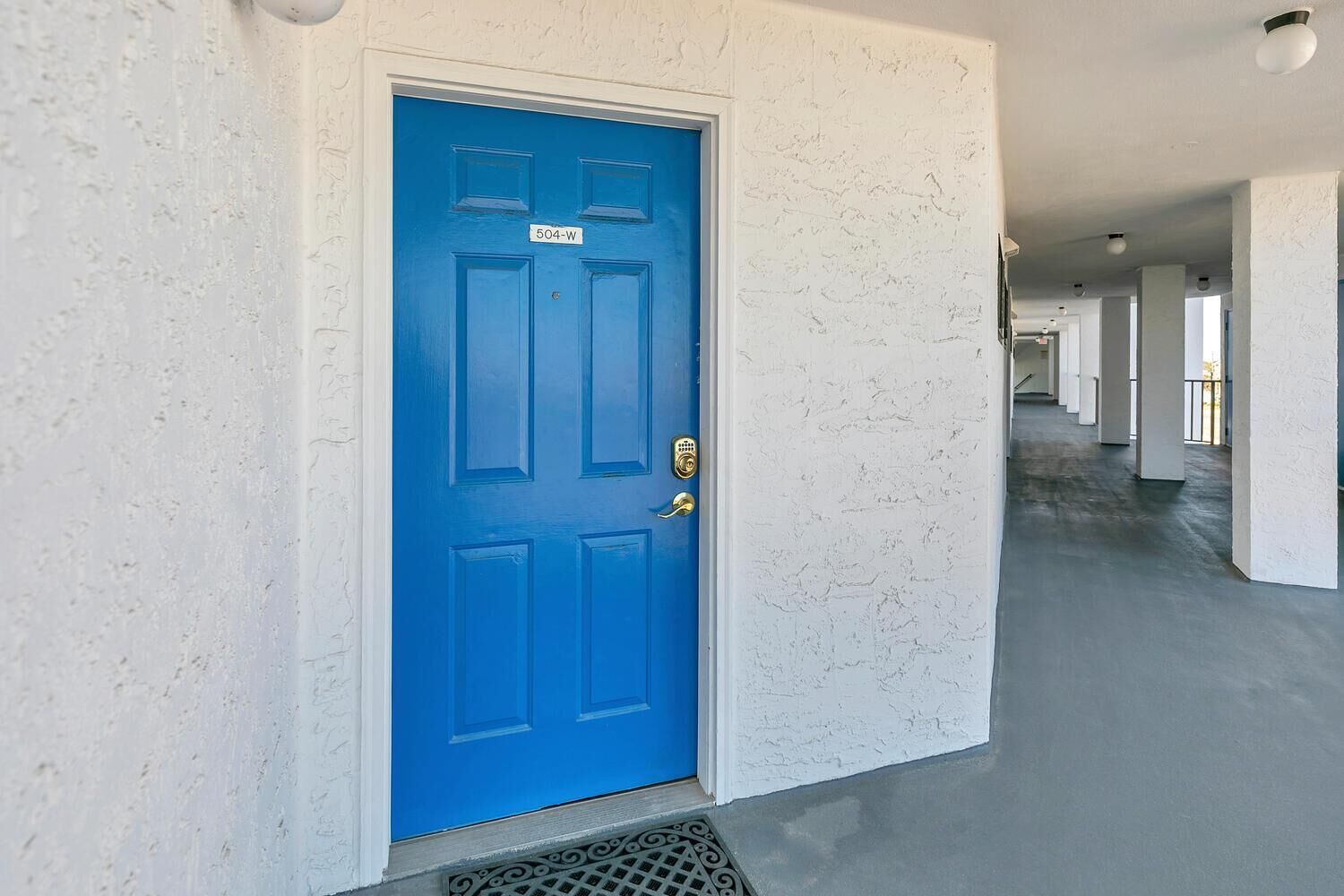 A blue door with a number plate, a white textured wall, and a concrete floor in a hallway.