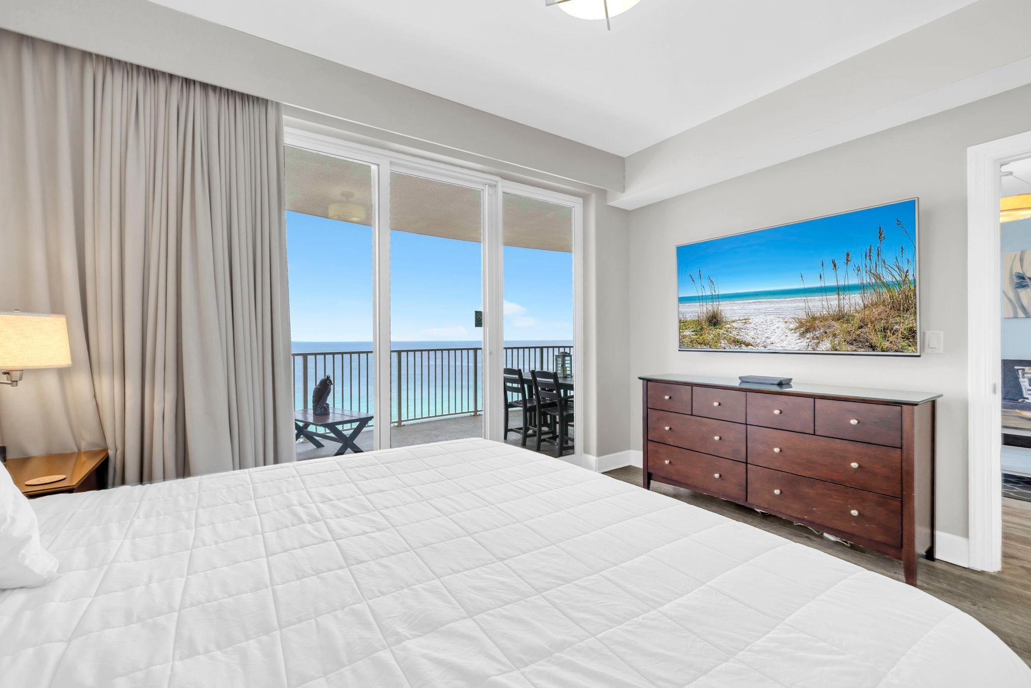 A hotel room with a large bed, a balcony with a view of the beach, a wooden dresser, and a painting of the beach.