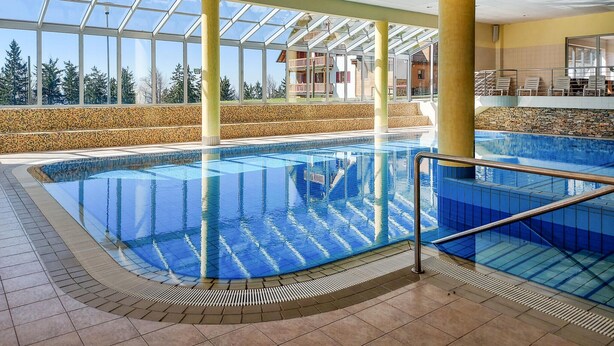Une grande piscine intérieure aux parois en verre transparent, dotée d’un sol carrelé et offrant une vue sur un immeuble avec balcon.