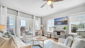 Ein geräumiges Wohnzimmer mit Deckenventilator, großen Fenstern mit Jalousien, Kamin und Blick ins Freie.