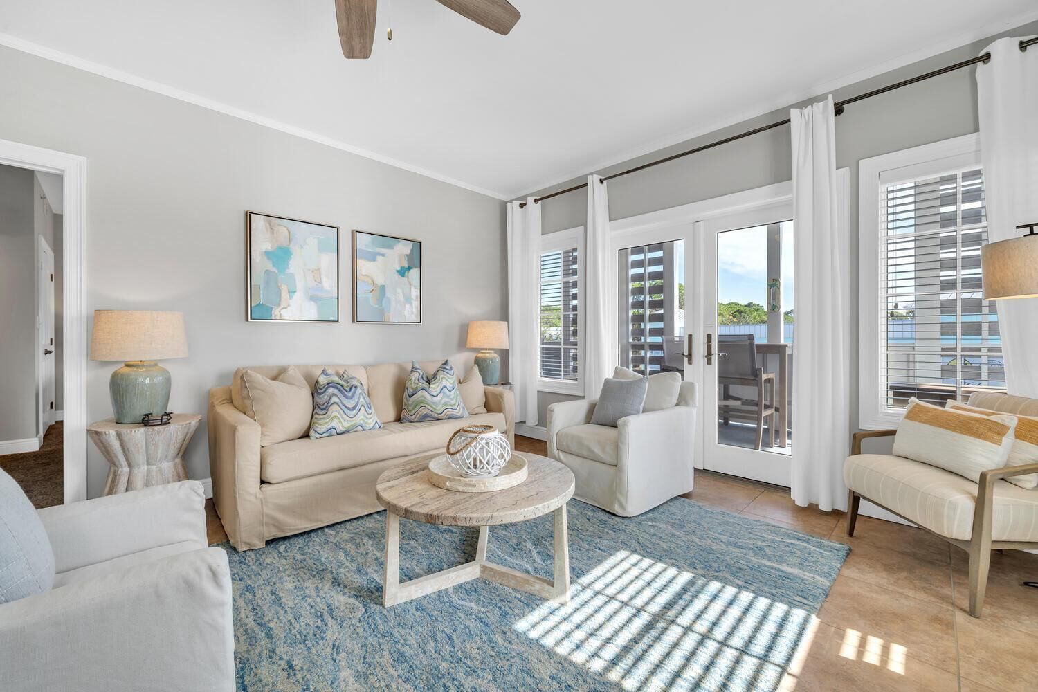 A spacious living room with a beige sofa, a round coffee table, and a blue rug. There are two side tables with lamps, a large window with blinds, and a ceiling fan.