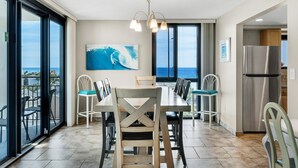 Un comedor con una mesa y sillas, un ventanal con vista al océano y una zona de cocina con refrigerador.