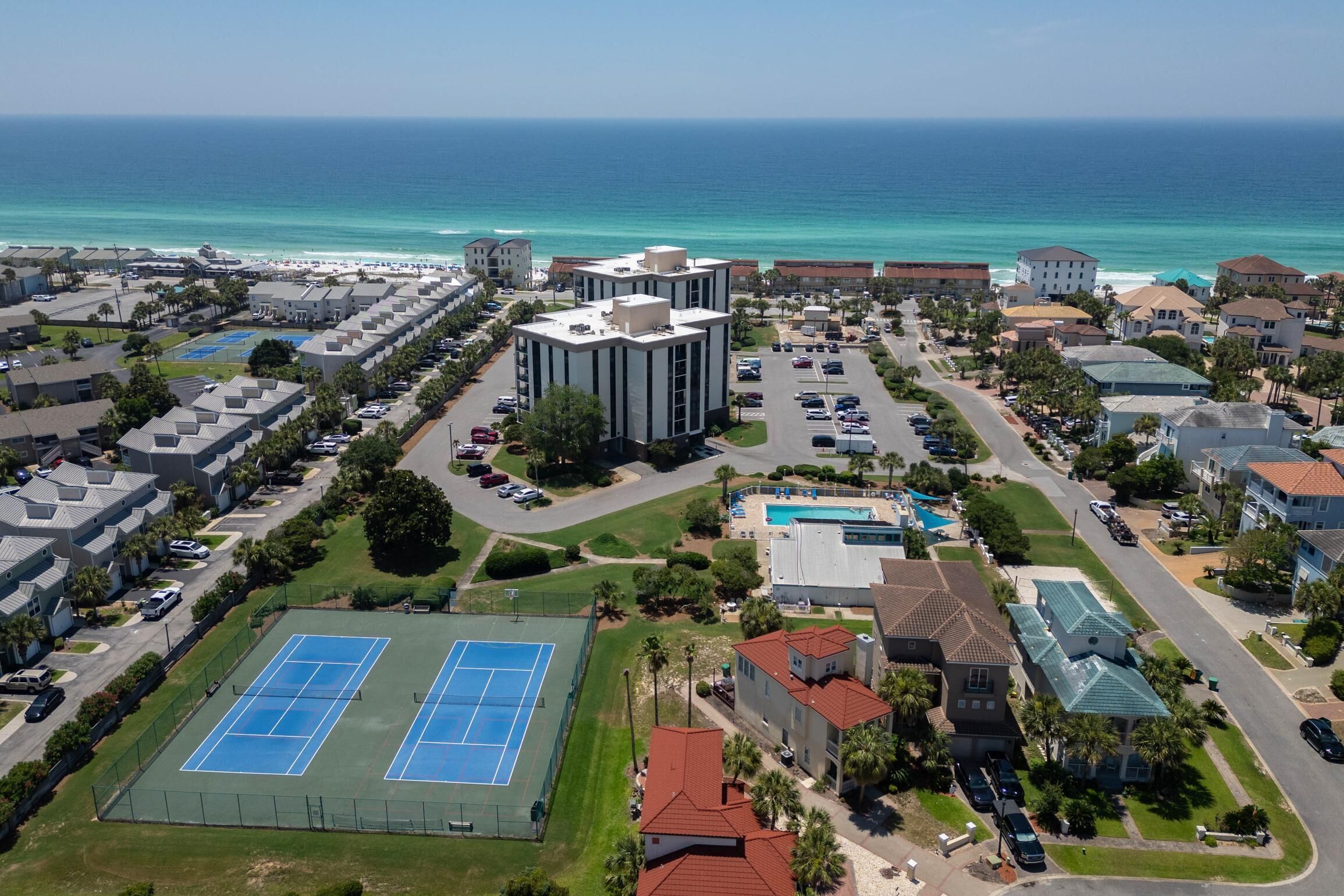 Vista aerea di una zona residenziale sulla costa, con un edificio grande, campi da tennis e una piscina.