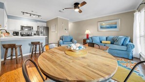 Une cuisine moderne avec des armoires blanches, un four à micro-ondes et un réfrigérateur. On y trouve un coin repas avec une table et des chaises en bois. Un ventilateur de plafond est visible au-dessus du coin repas.