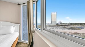 A bedroom with a bed, a window with curtains, and a view of a beach and buildings.