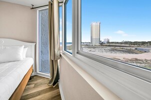 A bedroom with a bed, a window with curtains, and a view of a beach and buildings.