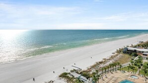 A beach view with a clear sky, a sandy beach, and a building with a pool.