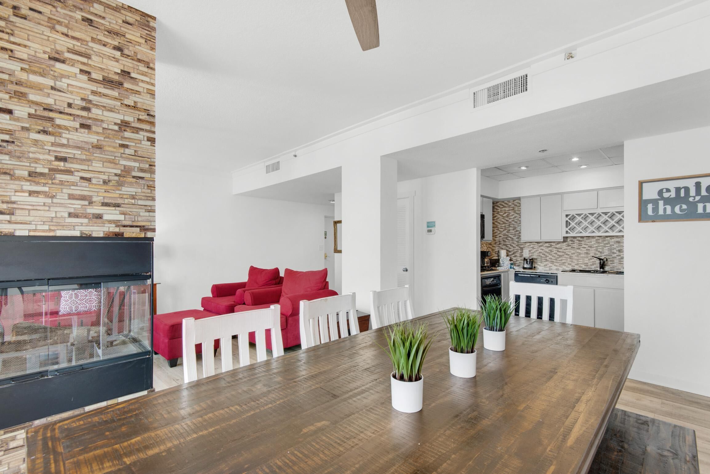 A modern living room with a wooden dining table, red sofa, and white chairs. A kitchen with white cabinets and a backsplash is visible in the background.