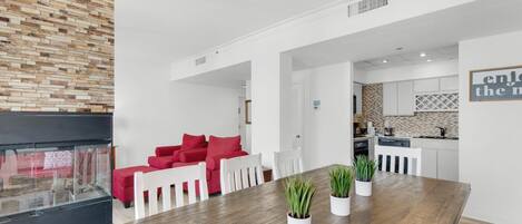 A modern living room with a wooden dining table, red sofa, and white chairs. A kitchen with white cabinets and a backsplash is visible in the background.