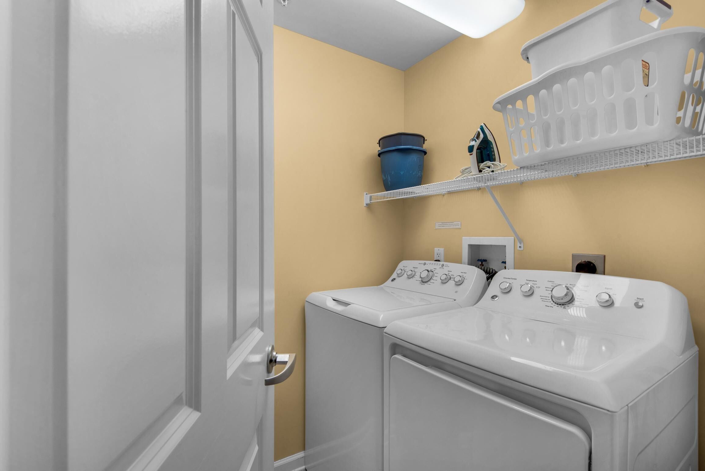 A laundry room with two white front-loading washers and dryers, a blue bucket, and a white laundry basket on a shelf.
