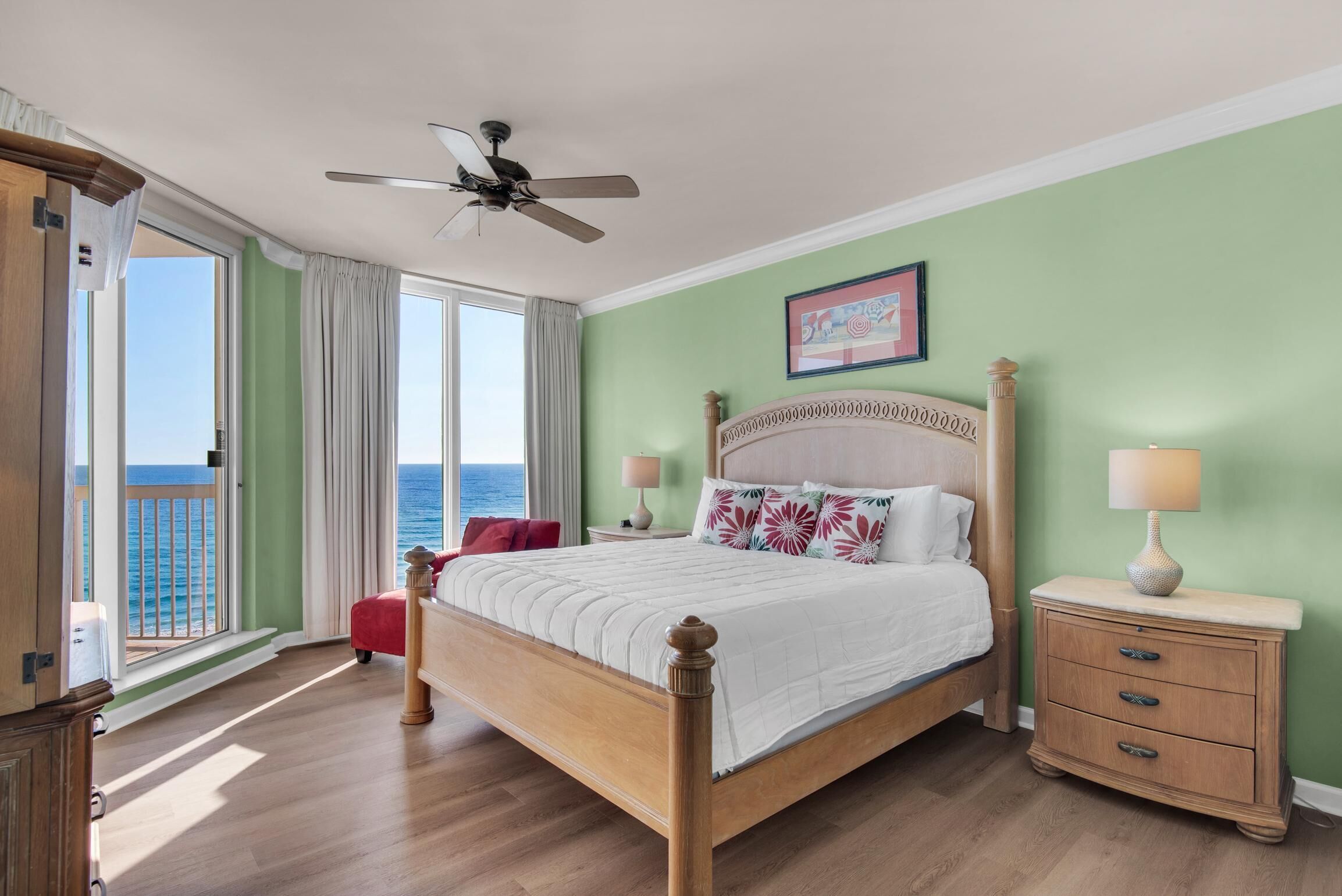 Une chambre à coucher avec un lit, une table de chevet, un ventilateur de plafond, une fenêtre avec des rideaux et une vue sur l’océan.