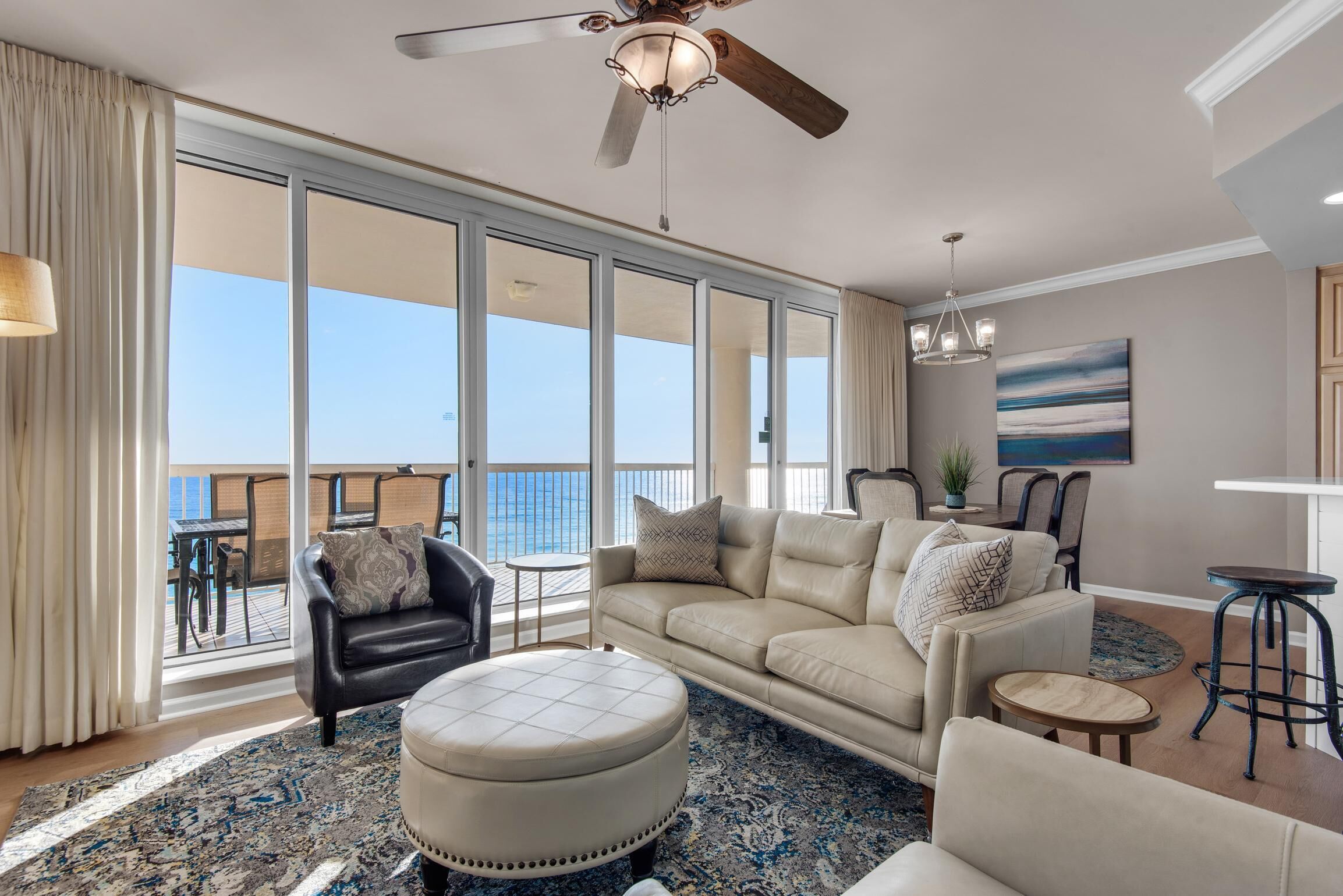 A spacious living room with a large window overlooking the sea, a ceiling fan, and a modern chandelier.