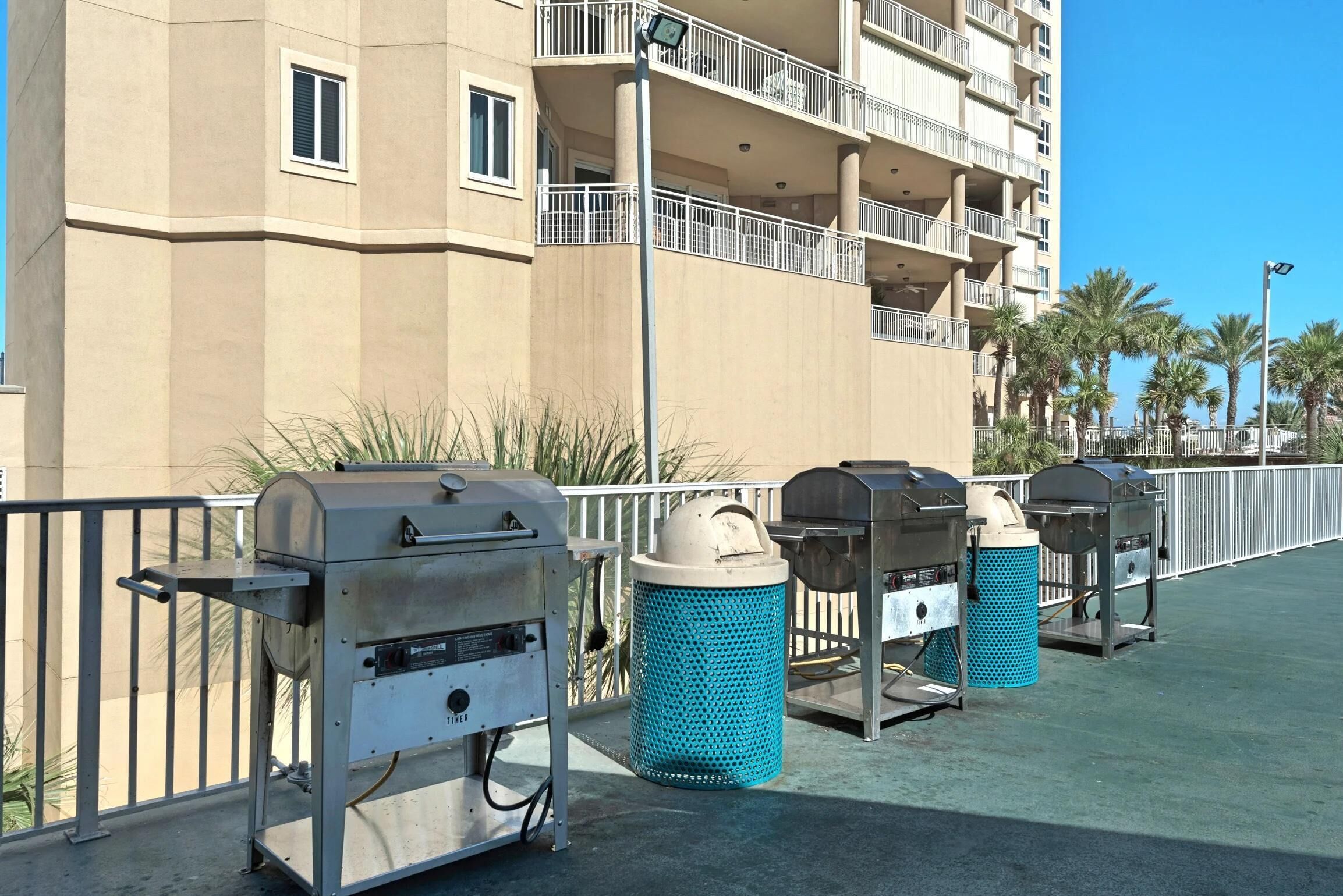 Outdoor barbecue grills and trash bins on a balcony with a building in the background.