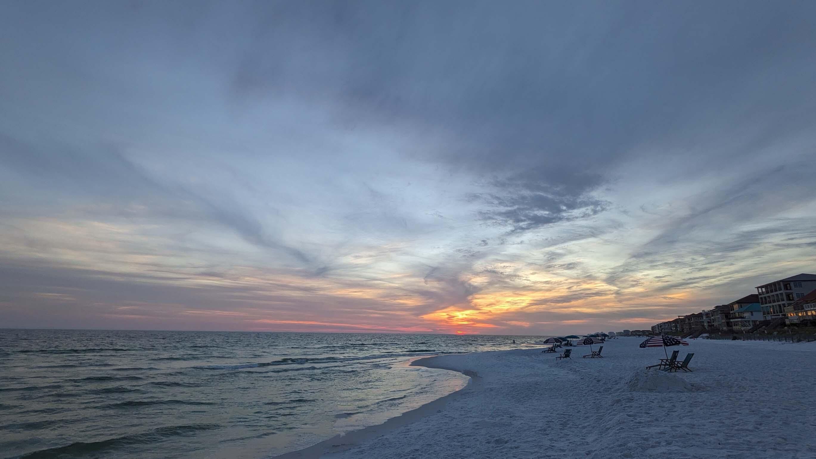 A beach with a sunset, calm sea, and beach chairs.