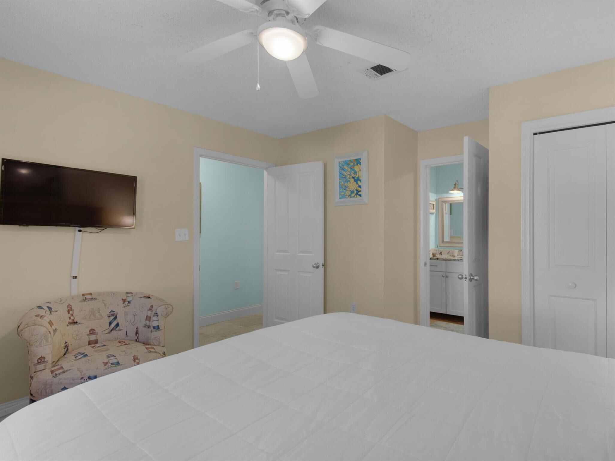 A bedroom with a bed, a television mounted on the wall, a ceiling fan, and a door leading to a bathroom.