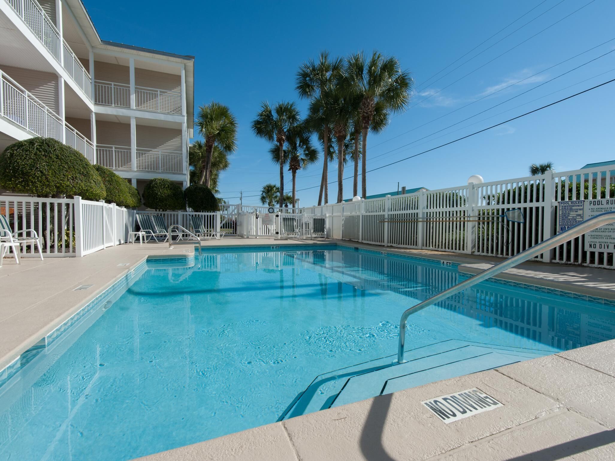 A swimming pool with a white fence, palm trees, and a multi-story building with balconies.