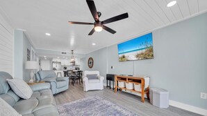 A spacious living room with a grey sectional sofa, a dining area with a table and chairs, a ceiling fan, and a large beach-themed painting on the wall. - Dunes of Panama 203c (Panama City Beach)