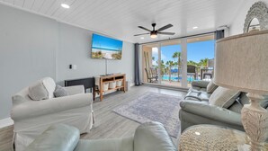 A modern living room with a flat-screen TV, a ceiling fan, and a view of a pool and palm trees through large sliding glass doors. - Dunes of Panama 203c (Panama City Beach)