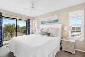 Une chambre avec un grand lit, un ventilateur de plafond, une table de chevet avec une lampe et une vue sur les palmiers, visible à travers une porte-fenêtre coulissante.