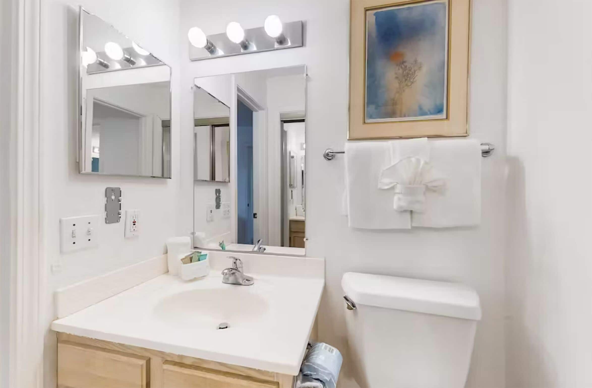 A bathroom with a white sink, two mirrors with lights, a toilet, and a towel rack.