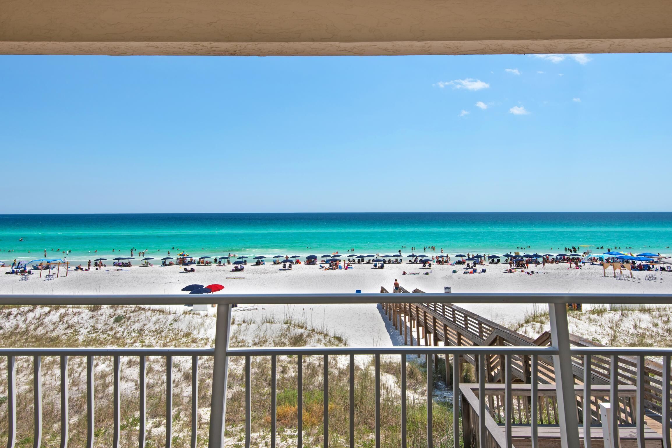 Blick auf einen Strand von einem Balkon mit Holzterrasse und blauem Himmel.