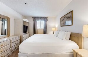 A hotel room with a large bed, a dresser, a television, a lamp, and a framed picture on the wall. - Crystal Sands 206b (Destin)