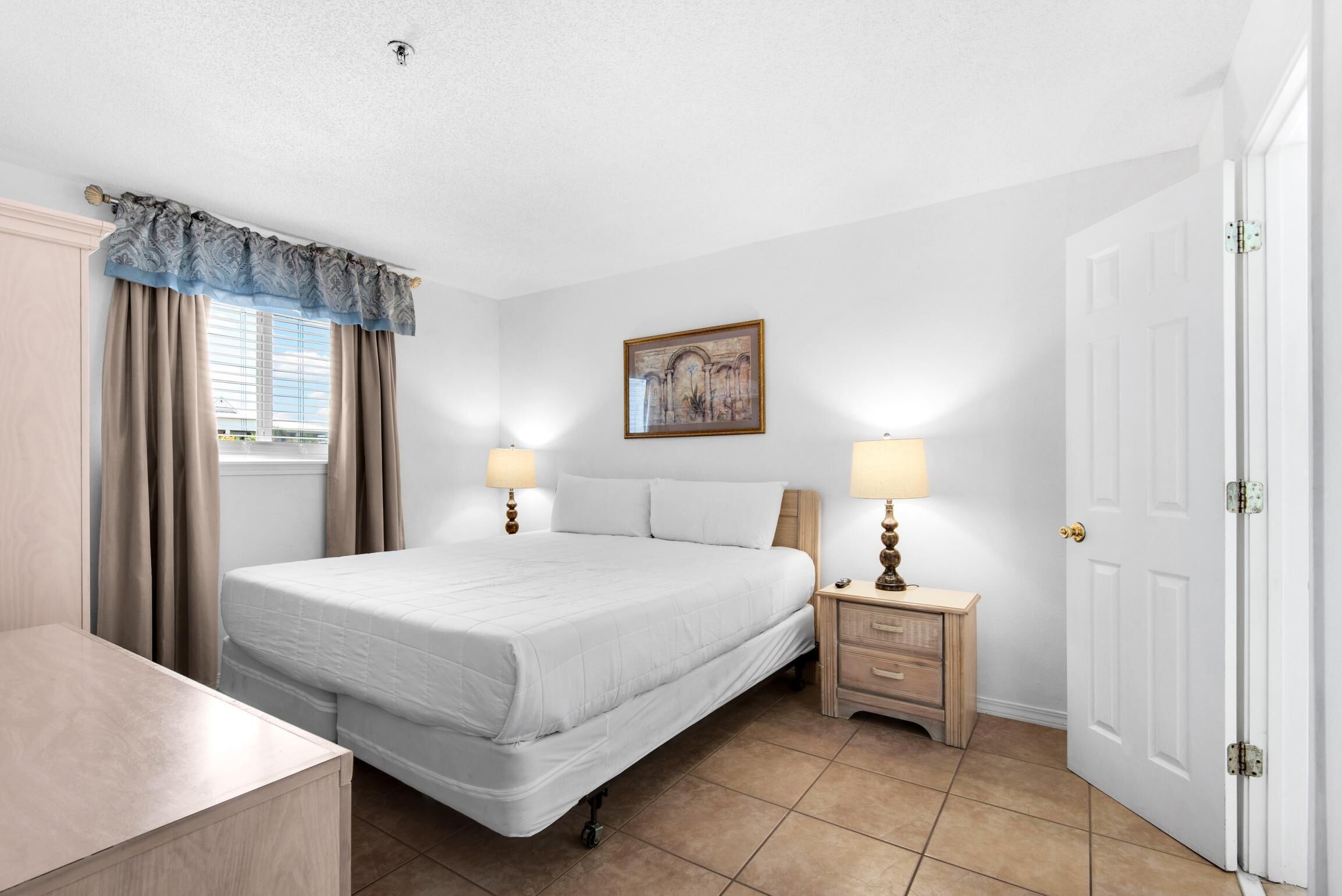 A bedroom with a bed, two bedside tables with lamps, a nightstand, a door, and a window with curtains.