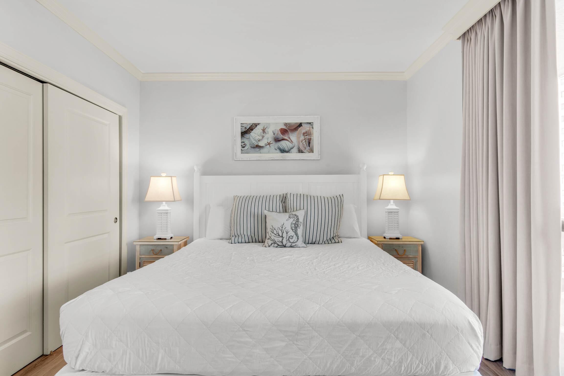 Un dormitorio ordenado con una cama grande, dos mesitas de noche con lámparas, un cuadro en la pared y una puerta a la izquierda.