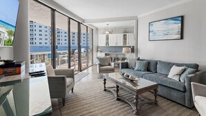 A spacious living room with a large window, a grey sofa, a coffee table, and a view of a beach and buildings. - Surfside Resort #208- Emerald Dreams (Miramar Beach)