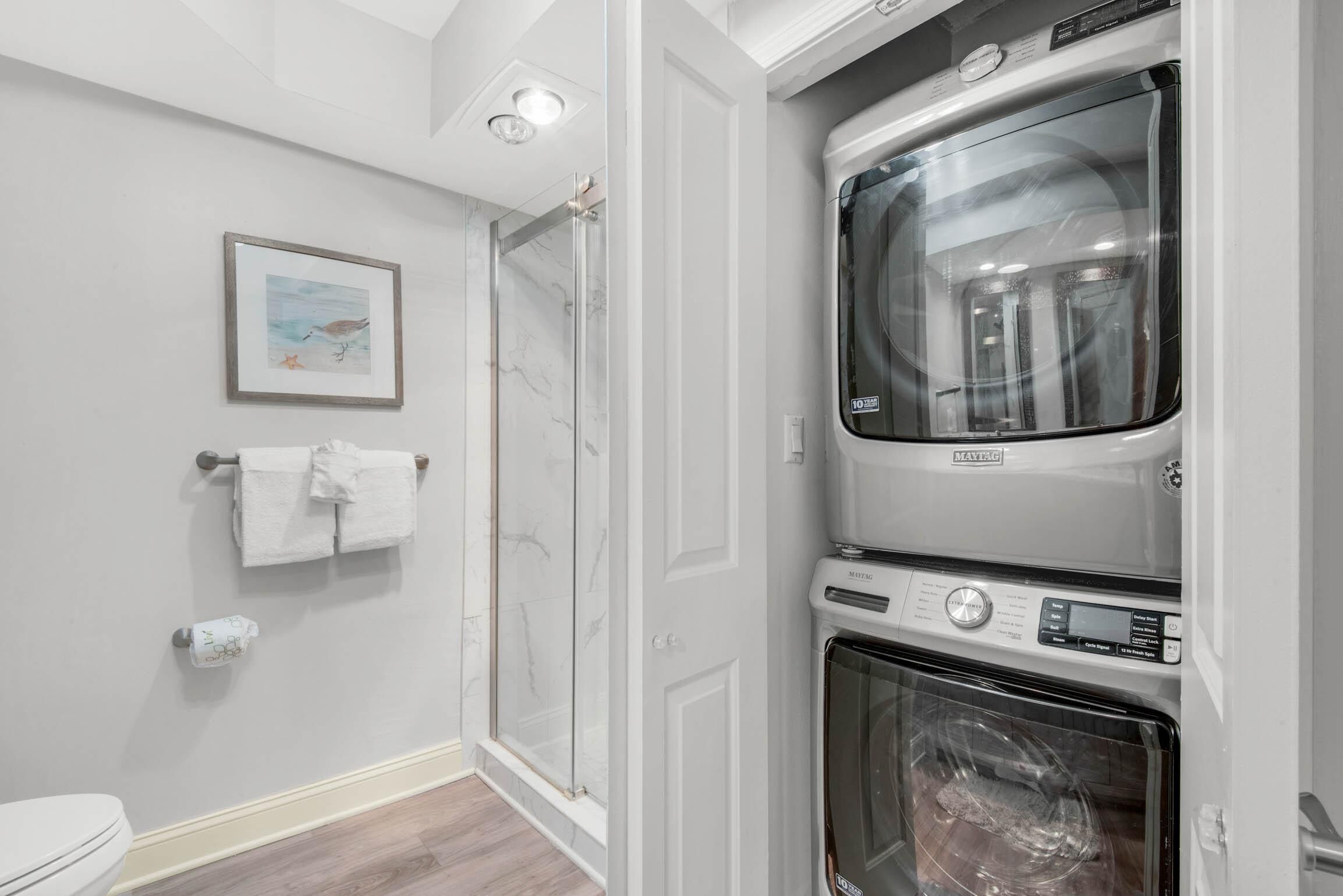 A bathroom with a glass shower, a toilet, a framed picture on the wall, and a stacked washer and dryer.