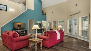 A spacious living room with two red sofas, a wooden coffee table, and a television displaying a sunset scene. - Captiva 67 (Miramar Beach)