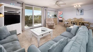 Un salon avec un canapé gris, une table basse blanche, un téléviseur posé sur un meuble TV blanc, un coin repas avec une table et des chaises, et un balcon avec vue.