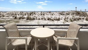 Un balcon avec deux chaises et une petite table, donnant sur une plage de sable avec une végétation clairsemée et un ciel dégagé.