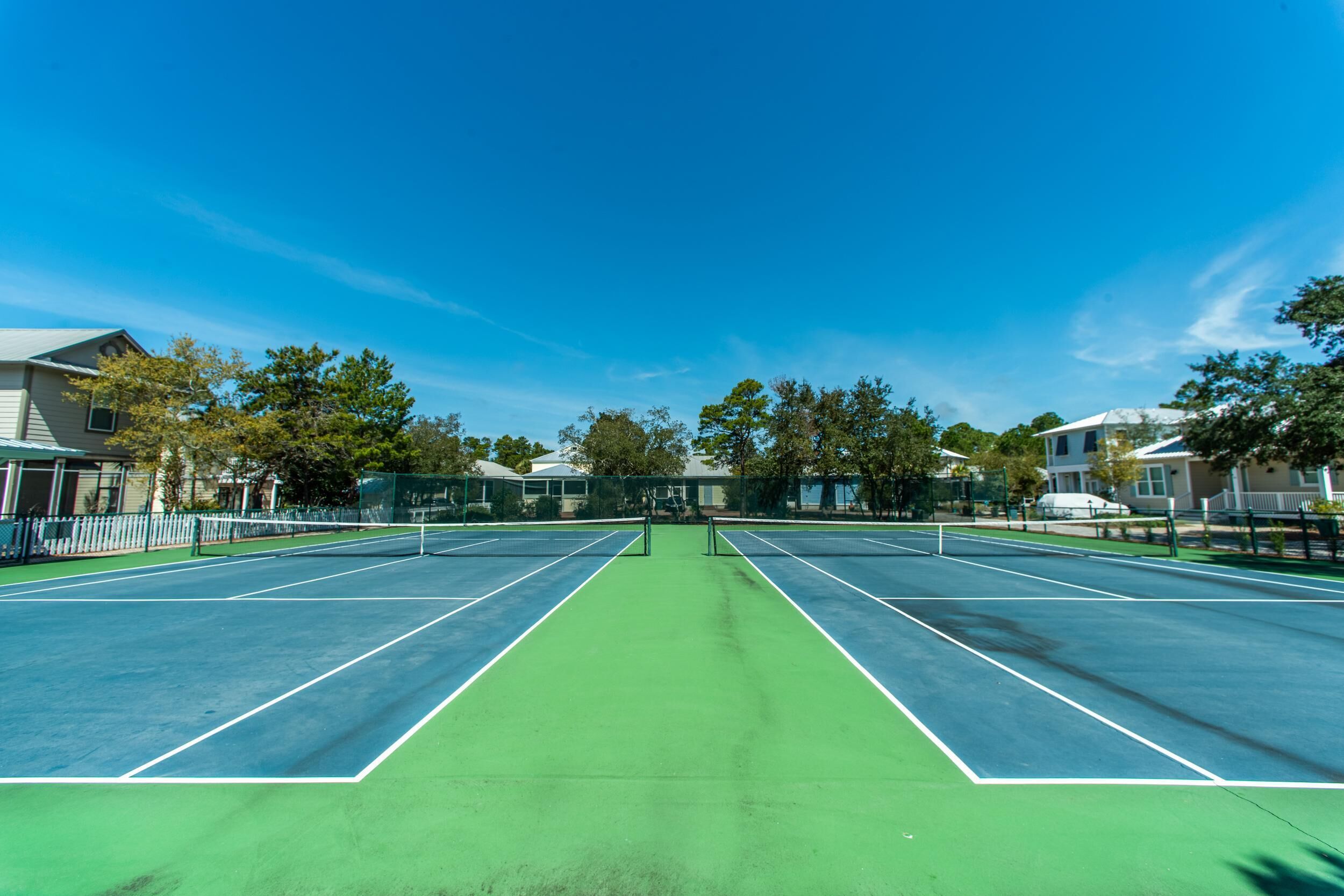 A tennis court with two courts, surrounded by trees and residential buildings.