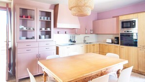 Une cuisine avec une table en bois, des chaises blanches, des armoires roses et des appareils électroménagers en acier inoxydable.