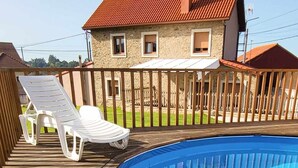 Une terrasse en bois avec une piscine, des chaises longues blanches et une maison en pierre avec un toit rouge.