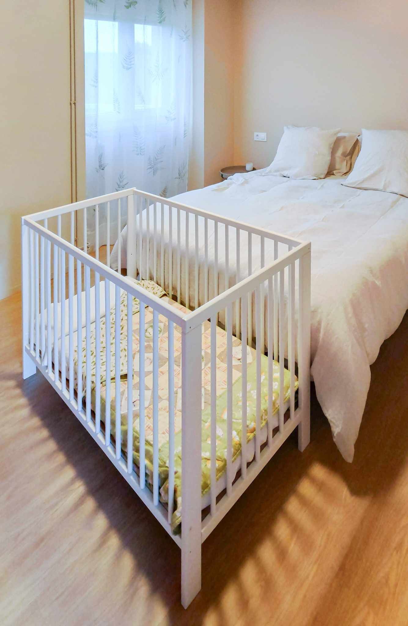A room with a white crib, a bed with white bedding, and a window with sheer curtains.