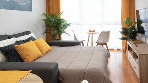 Moderni hotellihuone, jossa on suuri sänky, sohva, pieni pöytä ja televisio. Huoneessa on myös kasveja ja ikkuna, jossa on verhot.