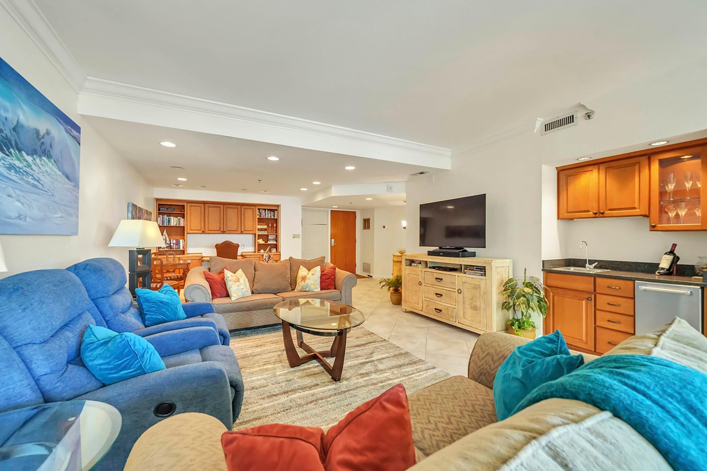 Un salón espacioso con televisor grande, asientos cómodos y una zona de cocina con armarios de madera y una mesa de comedor.